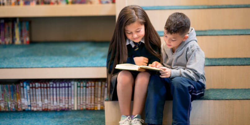 2 young students reading books together on the library stairs.